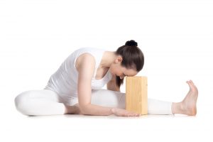 Sporty beautiful young beginning yoga student in white sportswear sitting in head to knee forward bend pose leaning on wooden block, doing Janu Sirsasana variation, studio full length isolated shot